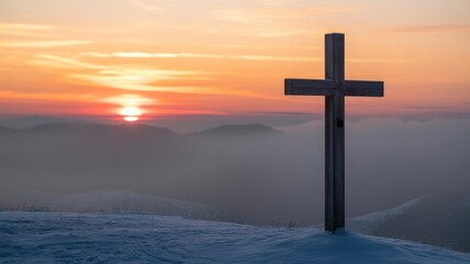A wooden cross stands majestically on a snow-covered mountain peak, silhouetted against a breathtaking sunset sky with hazy distant mountains.