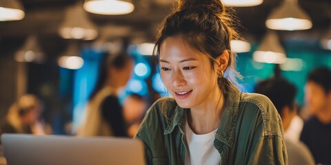 Smiling Asian office worker using laptop in modern co-working space with colleagues. Concept for business report, employee training and digital communication solutions