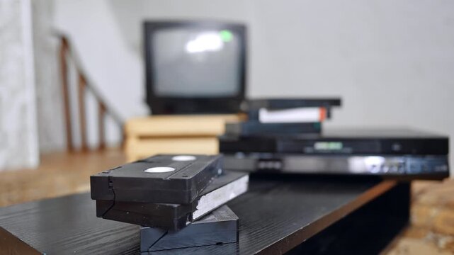 Old VHS cassettes and a player on the table. Hand presses the button on the recorder, takes a tape and puts on the pile. Close up.