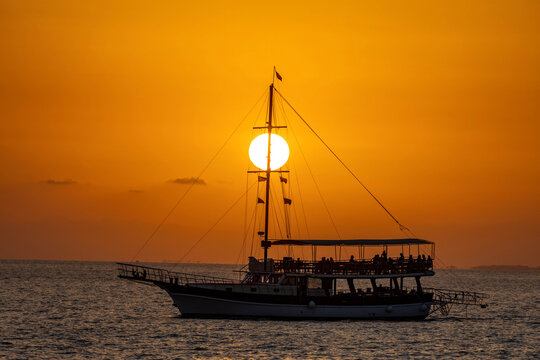 A tourist sailboat sails against the sunset with the orange sun disk perfectly aligned with its mast, creating a stunning minimalist silhouette scene on the Mediterranean Sea. Antalya, Turkey.