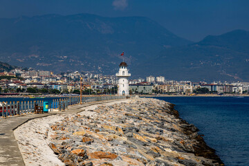 The iconic white lighthouse on Alanya's breakwater with Turkish flag, stretching toward the sea with city skyline and dark mountains in haze. Characteristic Mediterranean coastal scene.