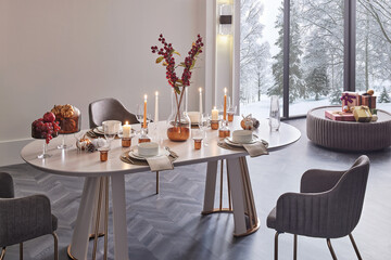 Close-up of a festive dining table set for Christmas. Cozy holiday ambiance with lit candles and a snowy winter window view.