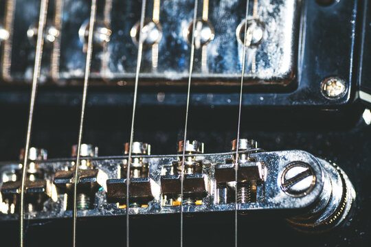 Reveals weathered bridge of electric guitar with taut strings stretching over brass saddles and adjustment screws. In background humbucker pickup adds to gritty, vintage allure under subtle lighting