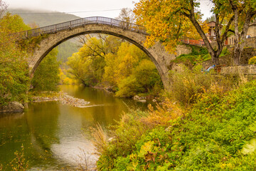 Iconic Single-Arch Stone Bridge of Vovousa Village Spanning the Aoos River, Surrounded by Golden Autumn Foliage in Pindos, Greece