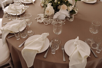 Fototapeta premium Elegant wedding reception table with white plates, folded napkins, glasses, and floral centerpiece on beige tablecloth. Concept for wedding decor, celebration, event planning, table setup.