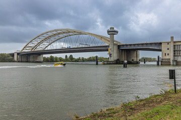 The imposing Van Brienenoord Bridge in Rotterdam arches over the Nieuwe Maas river, with a yellow water taxi speeding beneath the cloudy sky. Rotterdam, Netherlands. 19 October 2025.
