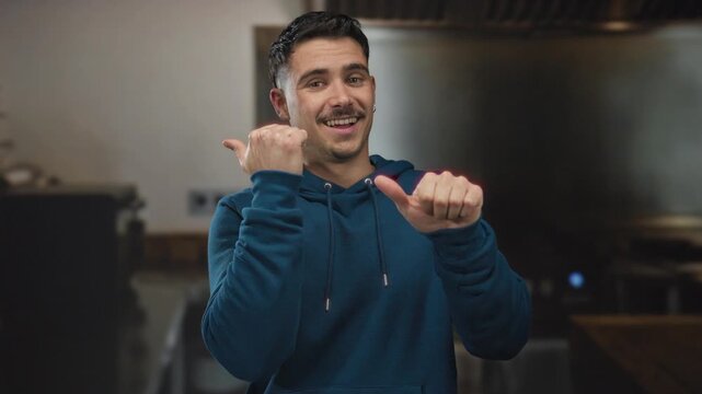 Young hispanic man in blue hoodie smiling and pointing thumbs sideways in kitchen setting offering vibrant indoor atmosphere and friendly gesture.