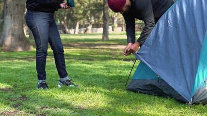 Two people working together to set up a camping tent on a grassy field, securing the structure to the ground during an outdoor trip in nature. - Powered by Adobe