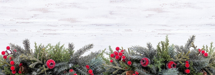 Christmas bottom border of snowy branches and red berry decorations. Top down view on a white wood banner background. Copy space.