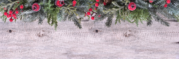 Christmas top border of snowy branches and red berry decorations. Above view on a light wood banner background. Copy space. © Jenifoto