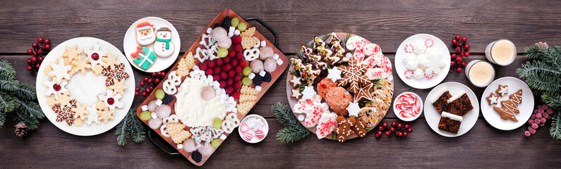 Christmas holiday cookies and sweets table scene. Above view on a dark wood banner background. Santa Claus grazing board, cookie platters and sweet plates.