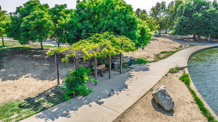 Elevated view of a serene park scene with a wooden pergola and benches. Lush green trees cast shadows on the sandy path alongside a calm lake. A peaceful outdoor setting ideal for relaxation or