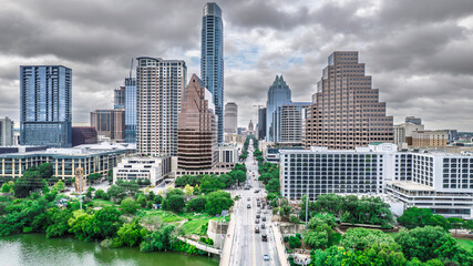 Panoramic aerial view of Austin, Texas skyline under dramatic cloudy skies. Skyscrapers dominate the cityscape alongside a busy street and lush green riverbanks. A vibrant urban scene showcasing
