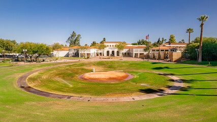 Panoramic view of a historic building in a desert park. A circular paved area leads to the stately structure under a clear blue sky. Palm trees and lush greenery complete this serene scene. Perfect