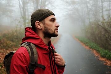 Side view of traveler with backpack gazing into foggy autumn forest road. Calm and thoughtful atmosphere