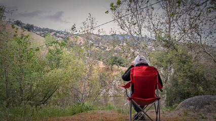 A lone backpacker sits in a folding chair overlooking a serene river valley. Moody landscape, reflection, contemplation, nature, peaceful moment.