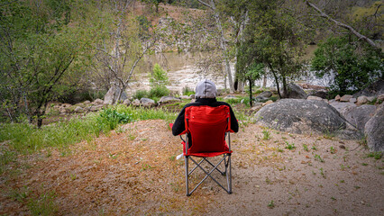 A lone backpacker sits in a folding chair overlooking a serene river valley. Moody landscape, reflection, contemplation, nature, peaceful moment.