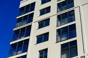 A fragment of the façade of a modern apartment building: white walls, large rectangular windows with dark frames, and balconies on the left. A bright blue sky appears in the background.