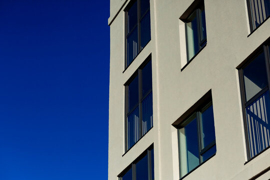 A fragment of the facade of a modern building with rectangular windows against a bright blue sky. Clean lines, light-colored walls, and a laconic architectural style.