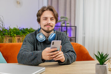 Caucasian man sits at table uses mobile phone smiles at home office room apartment. Young smiling guy texting share messages content on smartphone social media applications online watching relax movie