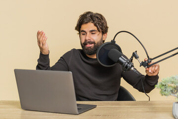 Happy blogger man recording podcast at table, giving workshop, webinar. Young Indian male speaking at microphone and laptop webcam, making video call. Arabian Hindu guy isolated on beige background