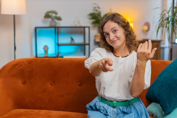 Hey you. Young woman smiling excitedly and pointing to camera, choosing lucky winner, indicating to awesome you, inviting, approve. Portrait of girl at home living room sitting on couch. Lifestyles.