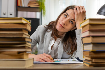 Exhausted Caucasian businesswoman looking at documents reports while working hard at home office desk. Stressed alone girl freelancer touching head in despair surrounded by stacks of books on desk