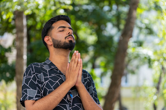 Bearded religion Indian man tourist praying with closed eyes to God asking for blessing, help, forgiveness outdoor. Young guy clasping hands wishing luck in urban sunny city street. Town lifestyle.