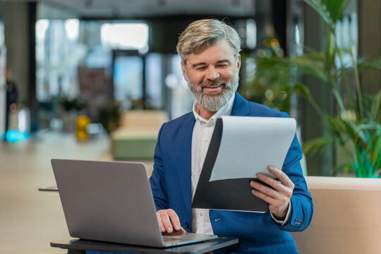 Happy businessman using laptop and working with documents, analyzing information, reading papers prepare financial report. Male executive in suit closing netbook after finishing work in office lobby.