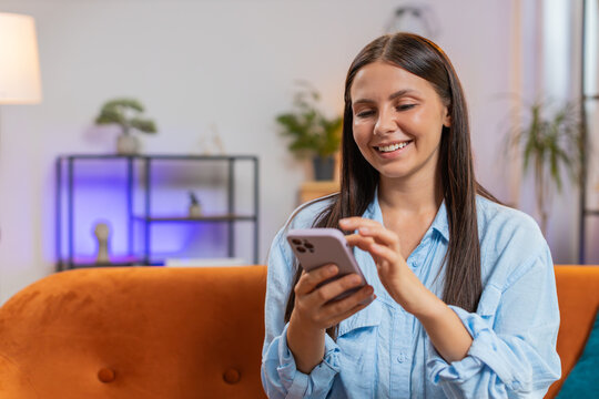 Happy young woman in casual clothes thinking and text messaging on smartphone social media app. Caucasian girl sitting on sofa and using mobile phone in living room at home. Lady watching online movie - Powered by Adobe