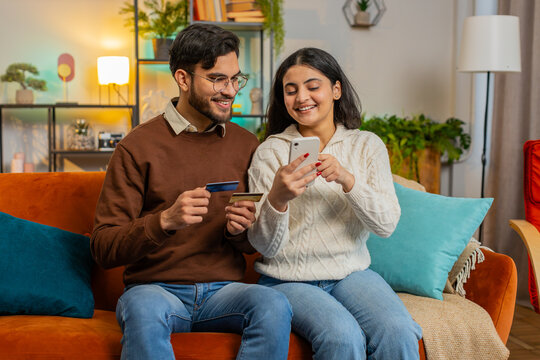 Happy Indian couple together doing online shopping by entering credit card number on smartphone while sitting on sofa at home. Smiling family are satisfied with a good promotion on goods in apartment.