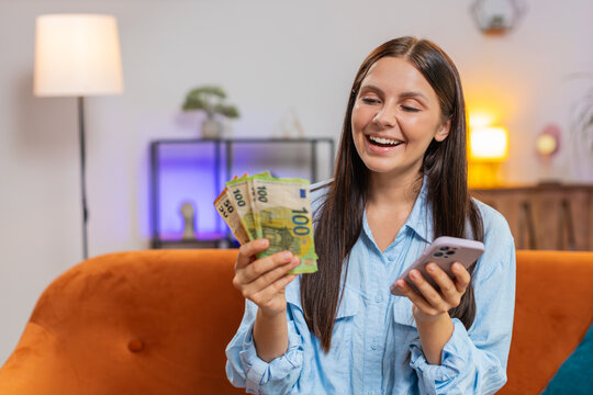 Smiling happy rich Caucasian woman counting money euro cash use smartphone calculate domestic bills sitting on home couch. Girl satisfied of income salary and saves money for planned vacation, gifts