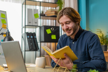 Caucasian young businessman reading interesting book, turning pages smiling enjoying literature, taking a rest after work. Happy cheerful freelancer guy with laptop relaxing at home office table
