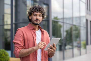 Close-up portrait of a young Indian man in a red shirt standing outside, holding a tablet and...