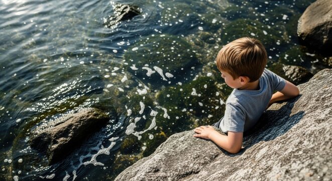 Young Caucasian boy sitting on a rock looking into the water. Childhood curiosity and summer exploration. Outdoor learning and environmental discovery. Peaceful moment of contemplation by the lake