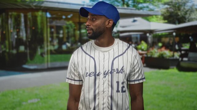 Man wearing baseball jersey and blue cap standing, looking to the side in front of a building; relaxed casual leisure.