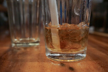 a glass of light brown drink with ice cubes and white straw on wooden table. The atmosphere looks warm and relaxed, possibly in a cafe or restaurant The main focus is on the front glass with a blurry 