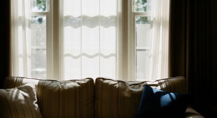 Bright sunlight through a sheer curtained window. Cozy living room sofa with pillows in deep shadow. Elegant home interior design and decor. Concept of quiet comfort and relaxation