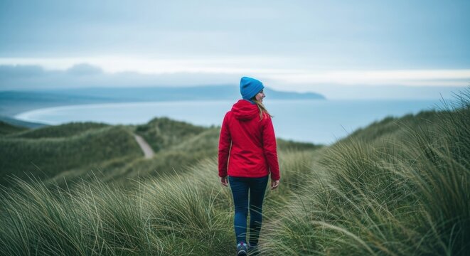 Caucasian female exploring a scenic coastal path. Outdoor adventure and travel lifestyle concept. Woman in red jacket walking through grassy dunes towards the ocean for wellness