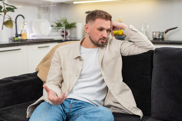 Young man at home looks down and feels exhausted after rough day and low mood and heavy stress. Caucasian guy on sofa breathes slow rubs neck and shows fatigue sadness and quiet pause for reset again
