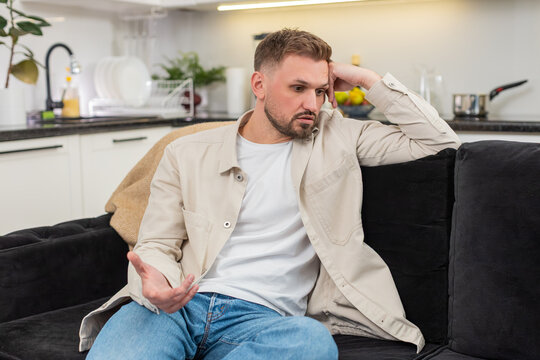 Young man at home looks down and feels exhausted after rough day and low mood and heavy stress. Caucasian guy on sofa breathes slow rubs neck and shows fatigue sadness and quiet pause for reset again