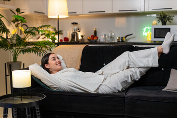 Young woman at night home lies back relaxes smiles enjoys quiet comfort takes breath slow. Caucasian girl on sofa at evening stretches legs feels cozy calm thanks climate control and silent appliance.