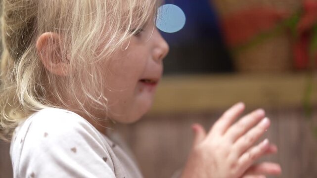 A young girl with blond hair eats a sloppy joe. She is eating with her hands at a table inside her home. It looks like she is enjoying her meal during a casual evening.