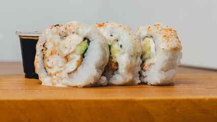 foto horizontal de 3 unidades de rollos acevichados, en primer plano, sobre una pequeña tabla de madera, acompañada de su salsa, sobre una mesa de madera.