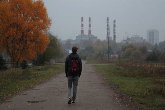 Young man with backpack walking on a quiet rural road towards distant industrial power plant chimneys in autumn