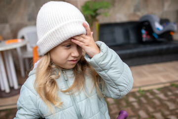 Fototapeta premium Upset young girl in a warm white knitted hat holding her forehead, looking stressed or disappointed on an outdoor patio