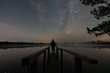 Silhouette of a man on a pier over a misty Estonian forest lake, watching the starry night sky and Milky Way.