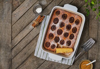 Cornmeal pie with fresh plums dusted with powdered sugar in a rectangular ceramic pan on a wooden background. Seasonal baking.