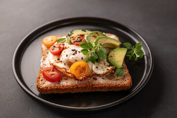Tasty homemade toast for breakfast with fried eggs, cream cheese, avocado, cherry tomatoes and fresh oregano leaves on black stone background close up.