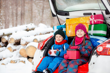 Merry Christmas concept. Two children in colorful winter suits open Christmas presents while sitting in trunk of white car surrounded by snow and pine trees.